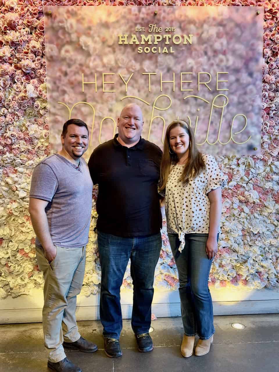 Three people stand smiling in front of a vibrant floral wall indoors, casually dressed, with "Hey There Nashville" elegantly written on it.