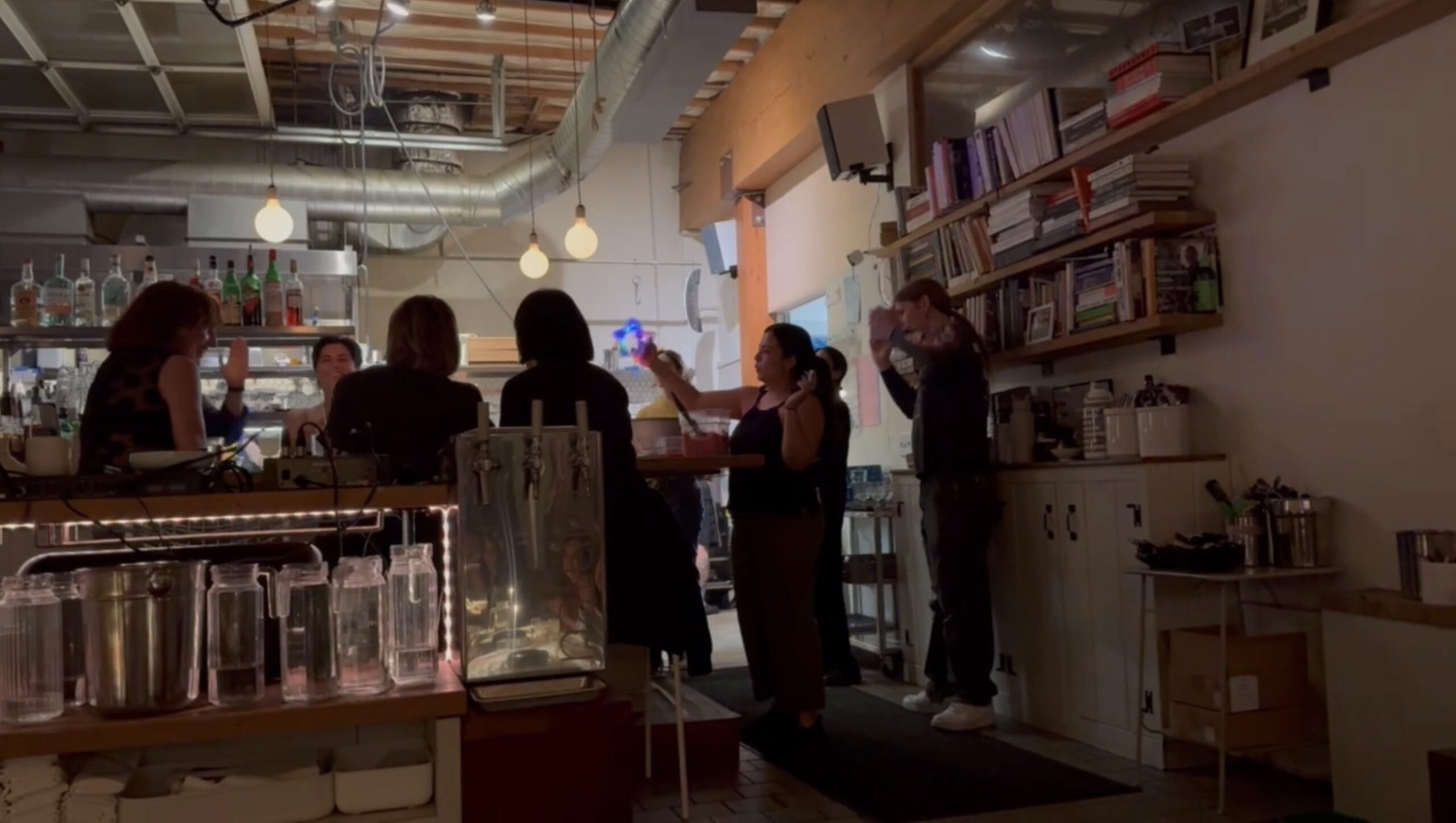 People are gathered inside a cozy bar or café with warm lighting, bookshelves, and a visible bar counter with drinks and glassware.