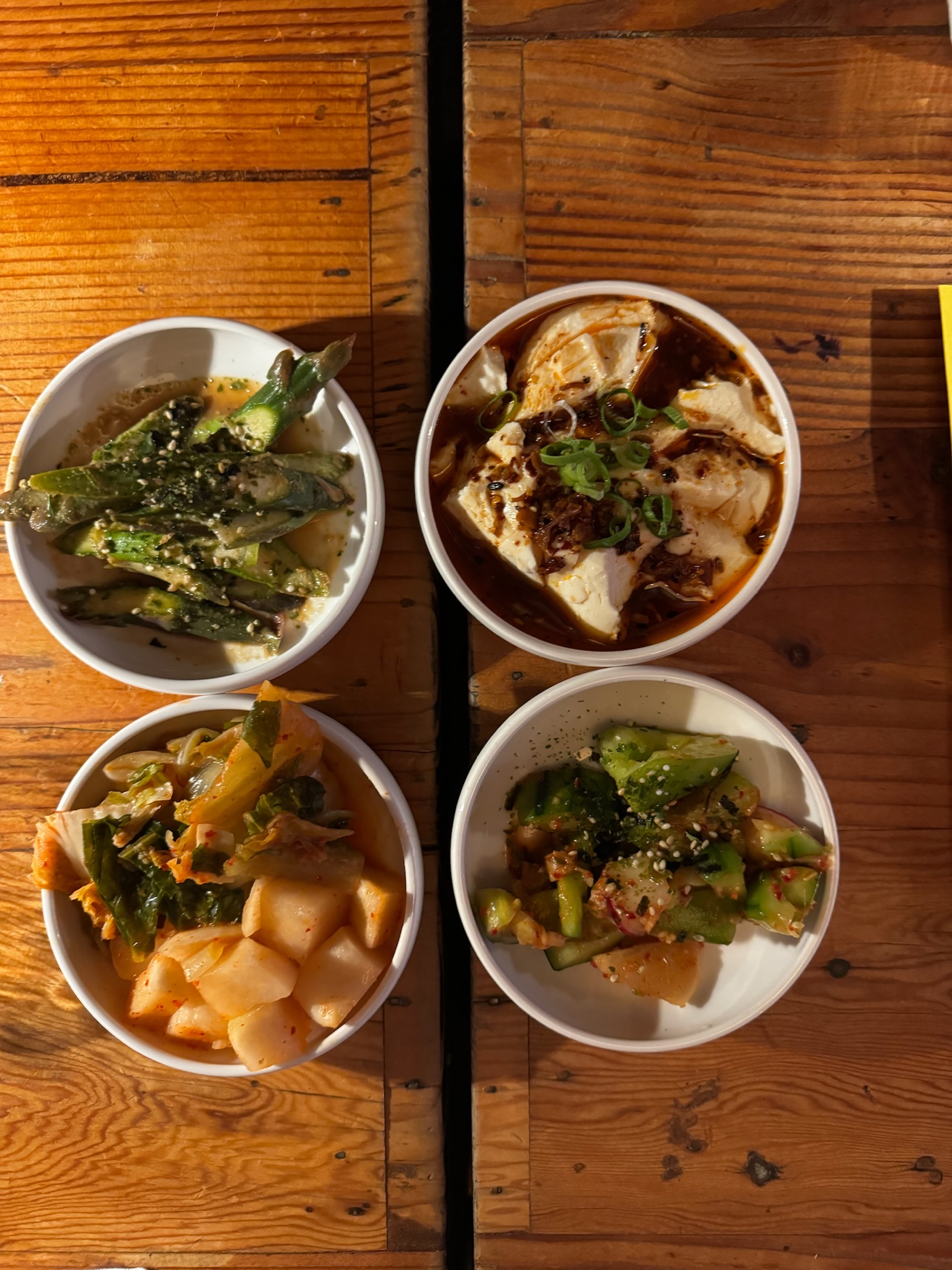 Four small bowls of food on a wooden table, containing tofu in sauce, sliced okra, kimchi, and cucumber salad with sesame seeds.