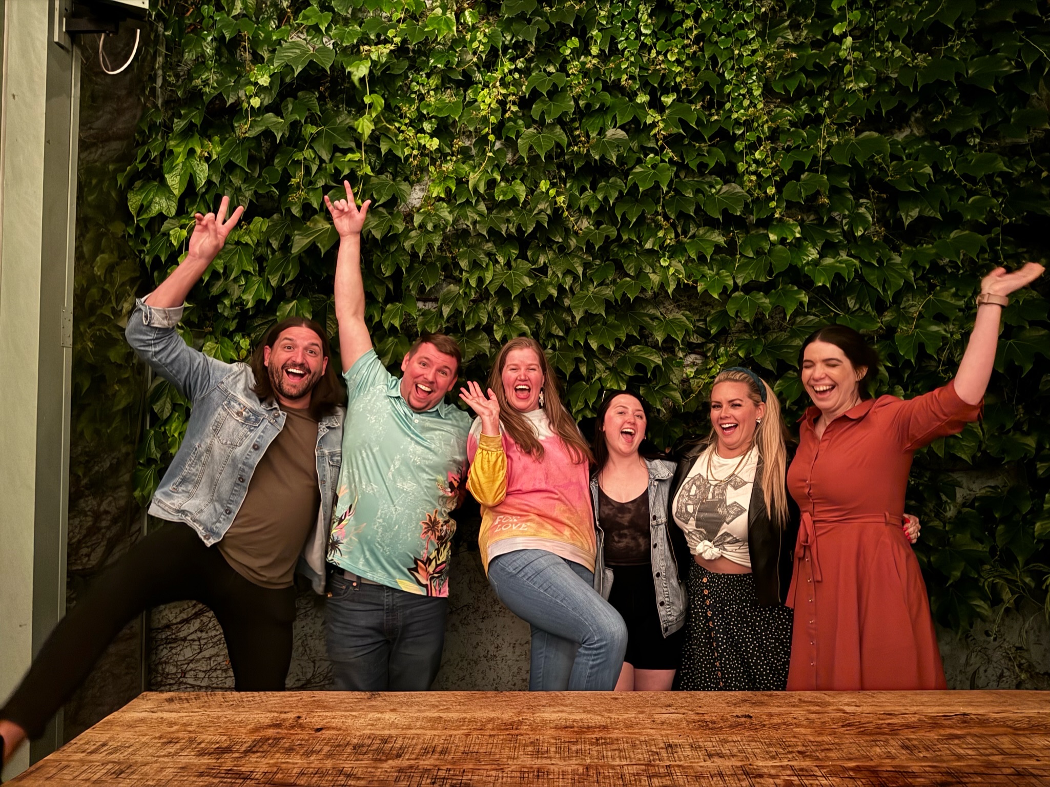 Six people stand in front of a leafy green wall, smiling and posing enthusiastically, with some raising their arms and one person lifting a leg. A wooden table is in the foreground.
