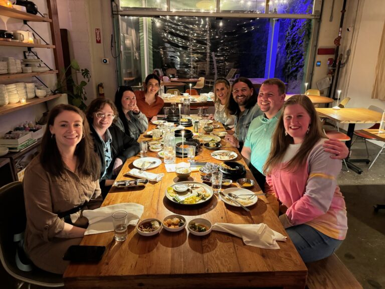 A group of eight people sit around a wooden table at a restaurant, sharing a meal with various dishes and drinks, smiling at the camera.