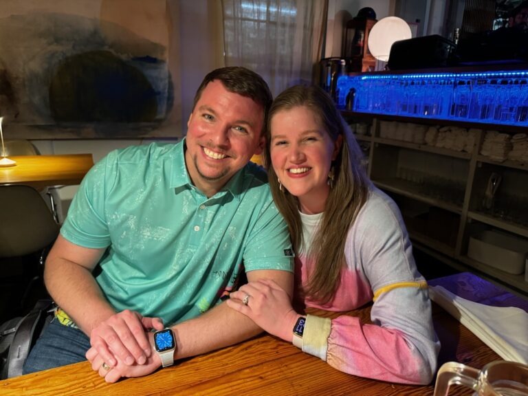 A man and a woman sit closely together at a wooden table in a restaurant, both smiling and wearing matching smartwatches.