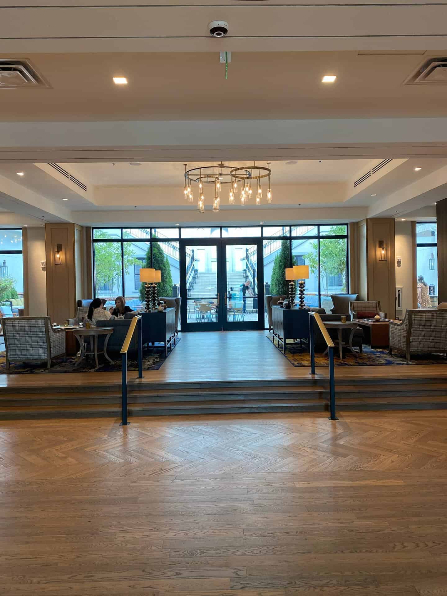 Spacious modern lobby with wooden floors, seating areas, and large windows overlooking Nashville. A chandelier hangs from the ceiling while people sit on the left, and outside stairs are visible through the bright windows.