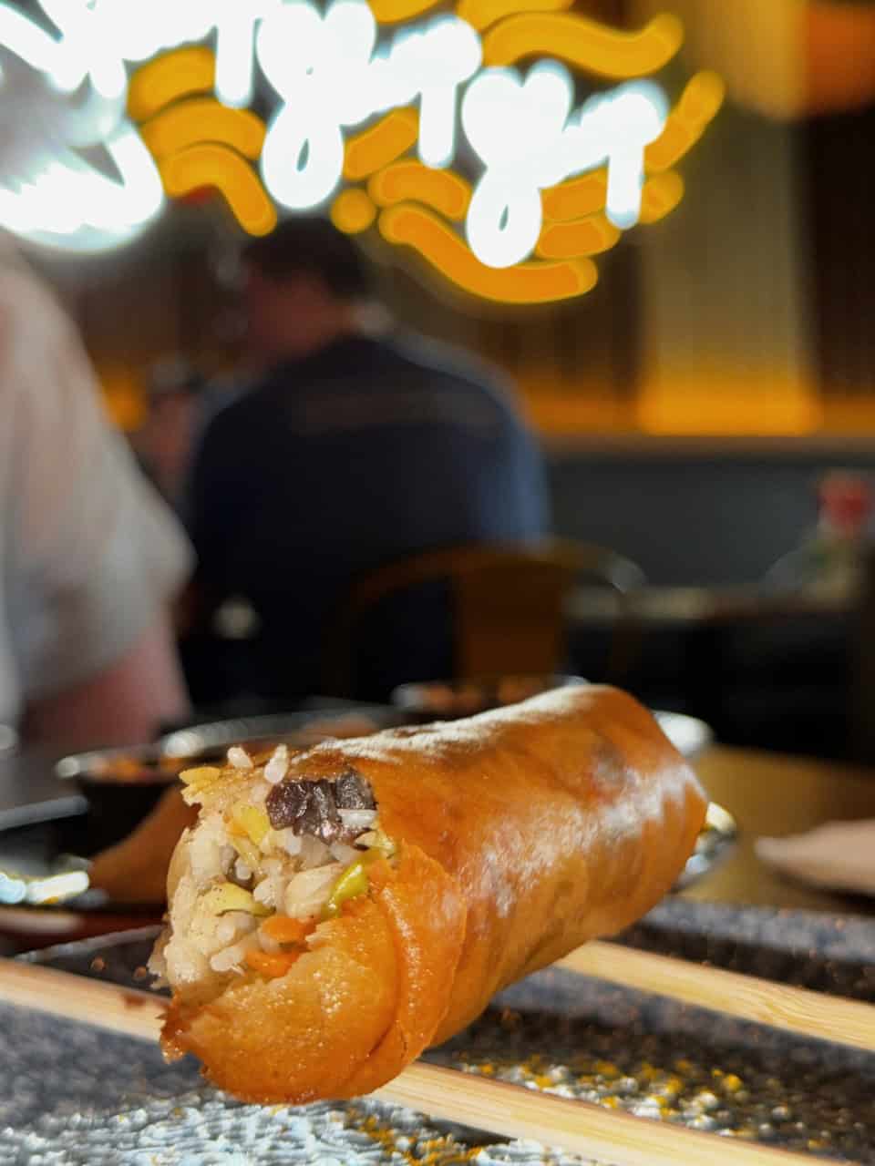 Close-up of a golden-brown spring roll filled with rice, vegetables, and meat, resting on wooden chopsticks. In the blurred background with warm lighting and neon signage, a hint of Nashville's vibrant energy adds to the scene.