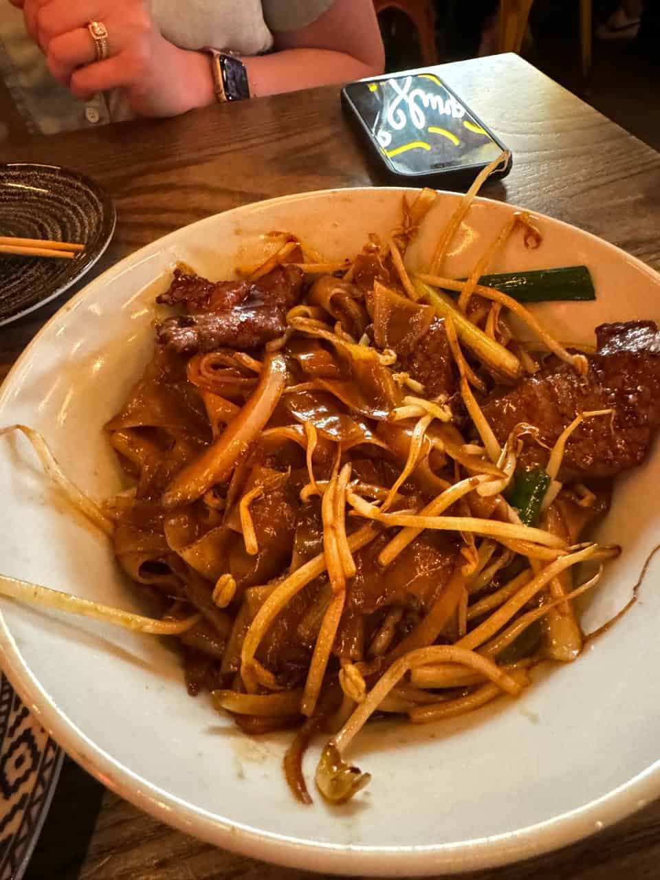A bowl of stir-fried noodles with beef, bean sprouts, and green onions sits invitingly on a wooden table. A person wearing a watch is seated nearby, perhaps planning their itinerary to explore Nashville, while their smartphone rests within easy reach.