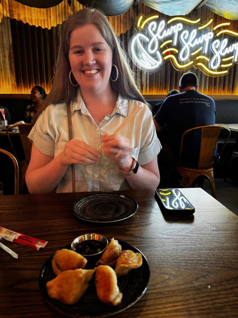 A person is savoring dumplings at a Nashville restaurant, with a neon "Slurp" sign glowing on the wall behind them.