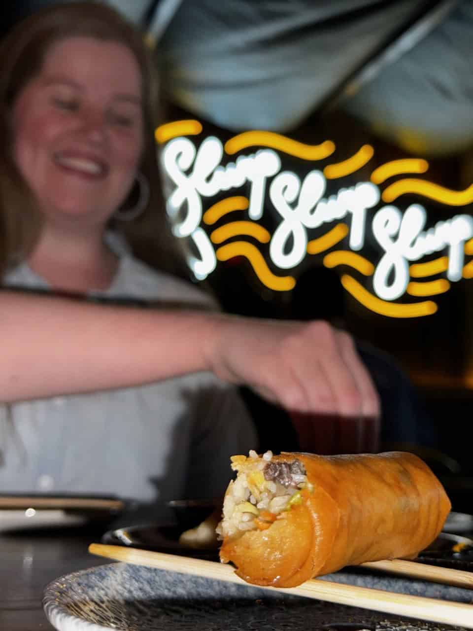 A person smiles while dining in a trendy Nashville eatery, with a spring roll on a plate in the foreground and neon "Slurp" signs glowing in the background.