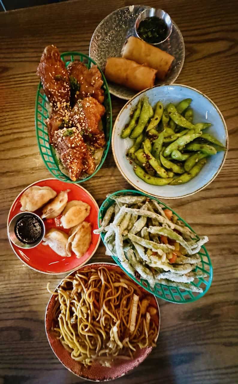 A table with Nashville-style fried chicken, spring rolls, edamame, dumplings, tempura vegetables, and a bowl of noodles.