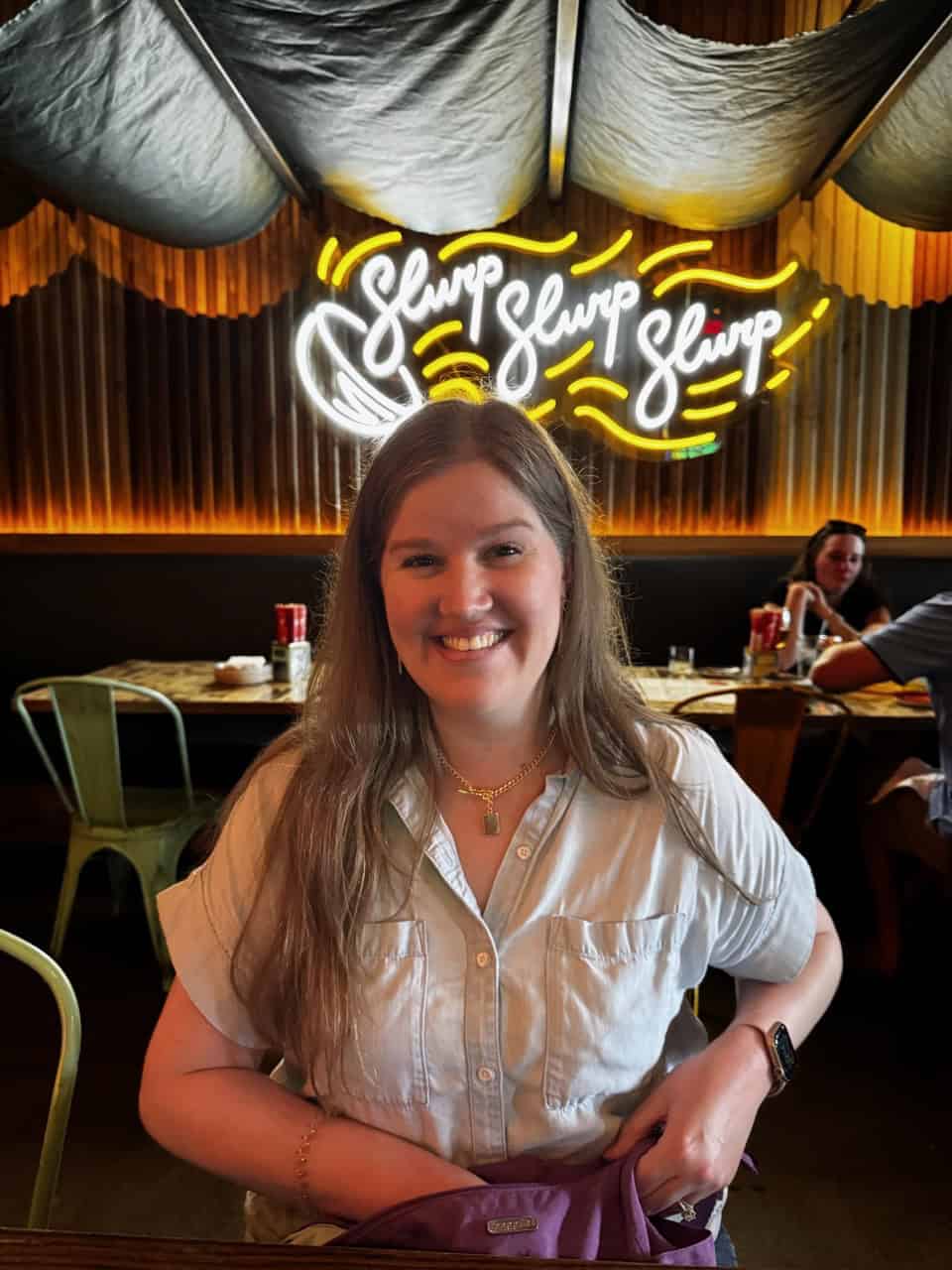 A person with long hair sits at a restaurant table in Nashville, smiling. A neon sign reading “Slurp Slurp Slurp” glows warmly in the background.