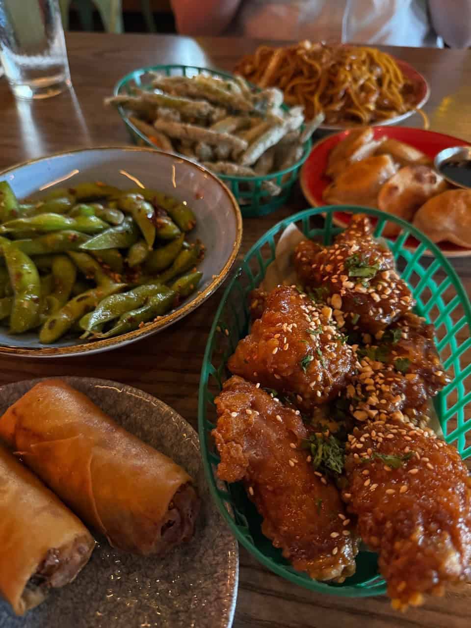 An array of Asian appetizers graces the table, featuring Nashville-inspired flavors in spring rolls, edamame, fried chicken wings, dumplings, vegetable tempura, and a delightful noodle dish.
