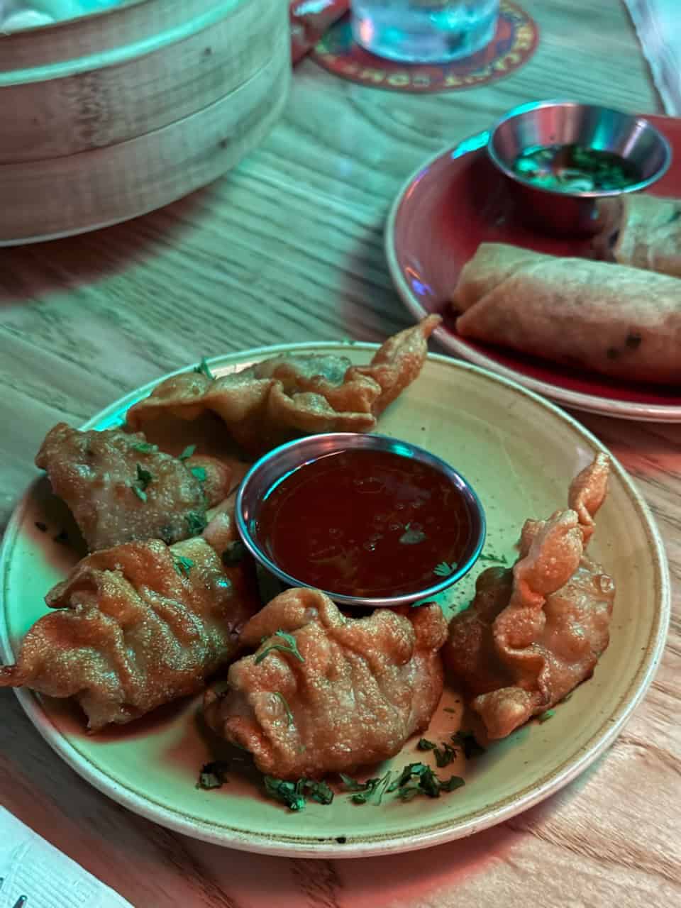 A plate of fried dumplings with a small bowl of red sauce in the center sits on a wooden table reminiscent of Nashville's rustic charm. Behind them are spring rolls and a bamboo steamer.
