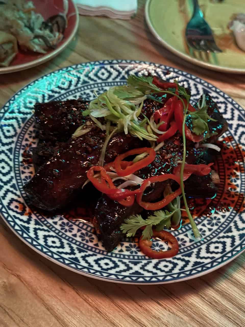 A decorative plate showcases Nashville-style grilled meat, garnished with sliced red chilies, cilantro, and green onions. It rests on a wooden table alongside other dishes partially visible.
