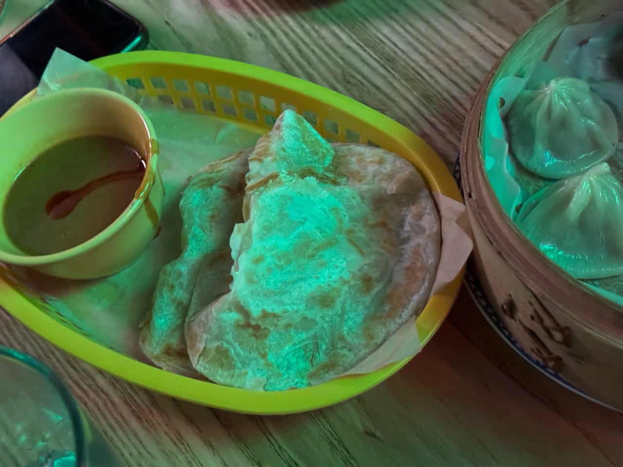 A basket with Nashville-inspired flatbreads and dipping sauce sits next to a steamer basket filled with dumplings on a wooden table.