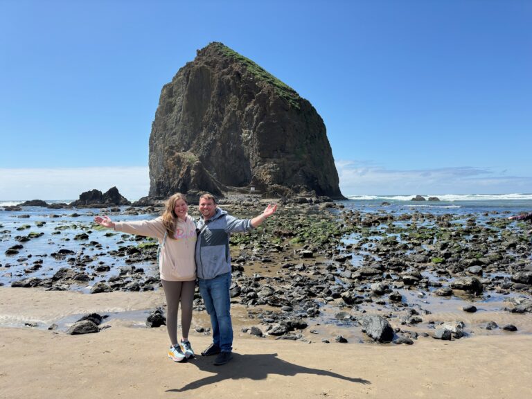 Two people stand smiling with arms outstretched on a rocky beach with a large sea stack and blue sky in the background.