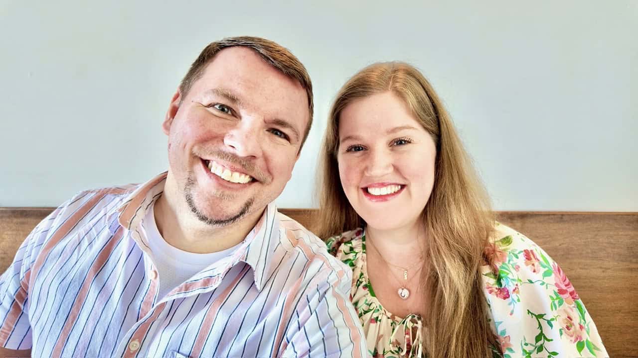 Two people are sitting closely together, smiling at the camera. The person on the left sports a striped shirt, while the one on the right is in a floral top. Their warmth and joy bring a touch of Nashville charm to an otherwise plain background.