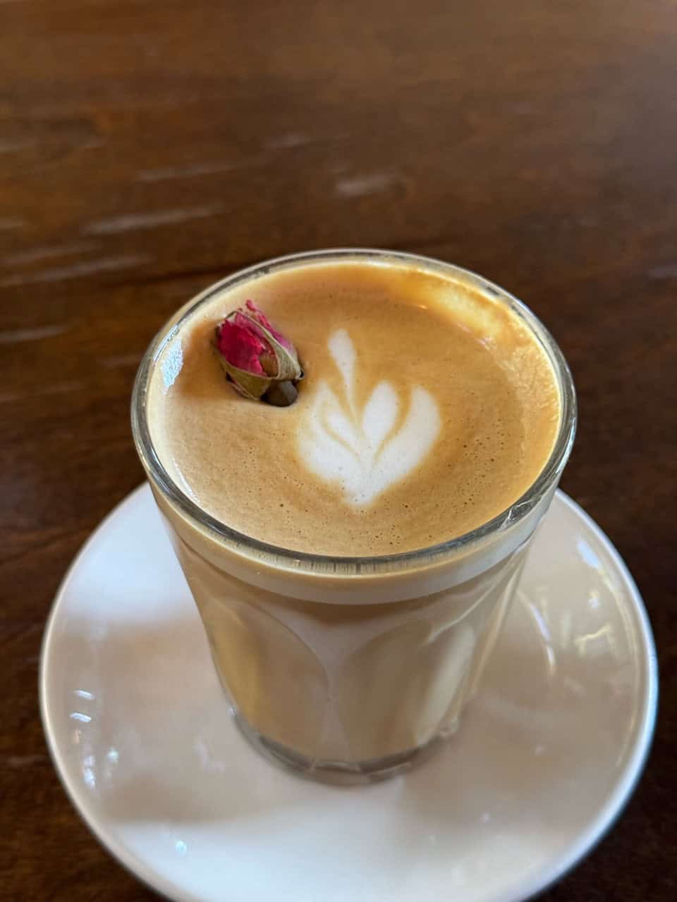 In Nashville's vibrant spirit, enjoy a glass of coffee adorned with leaf latte art and a delicate pink rosebud garnish, elegantly presented on a white saucer against a warm brown backdrop.