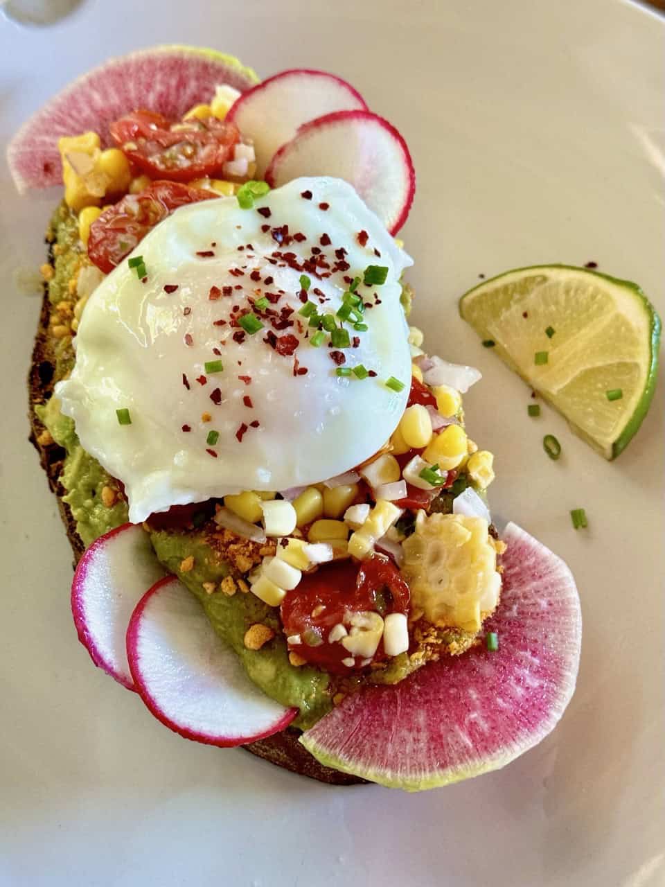 Nashville-inspired open-faced sandwich featuring avocado, corn, and tomatoes topped with a poached egg and radish slices, all beautifully arranged on a plate with a lime wedge garnish.