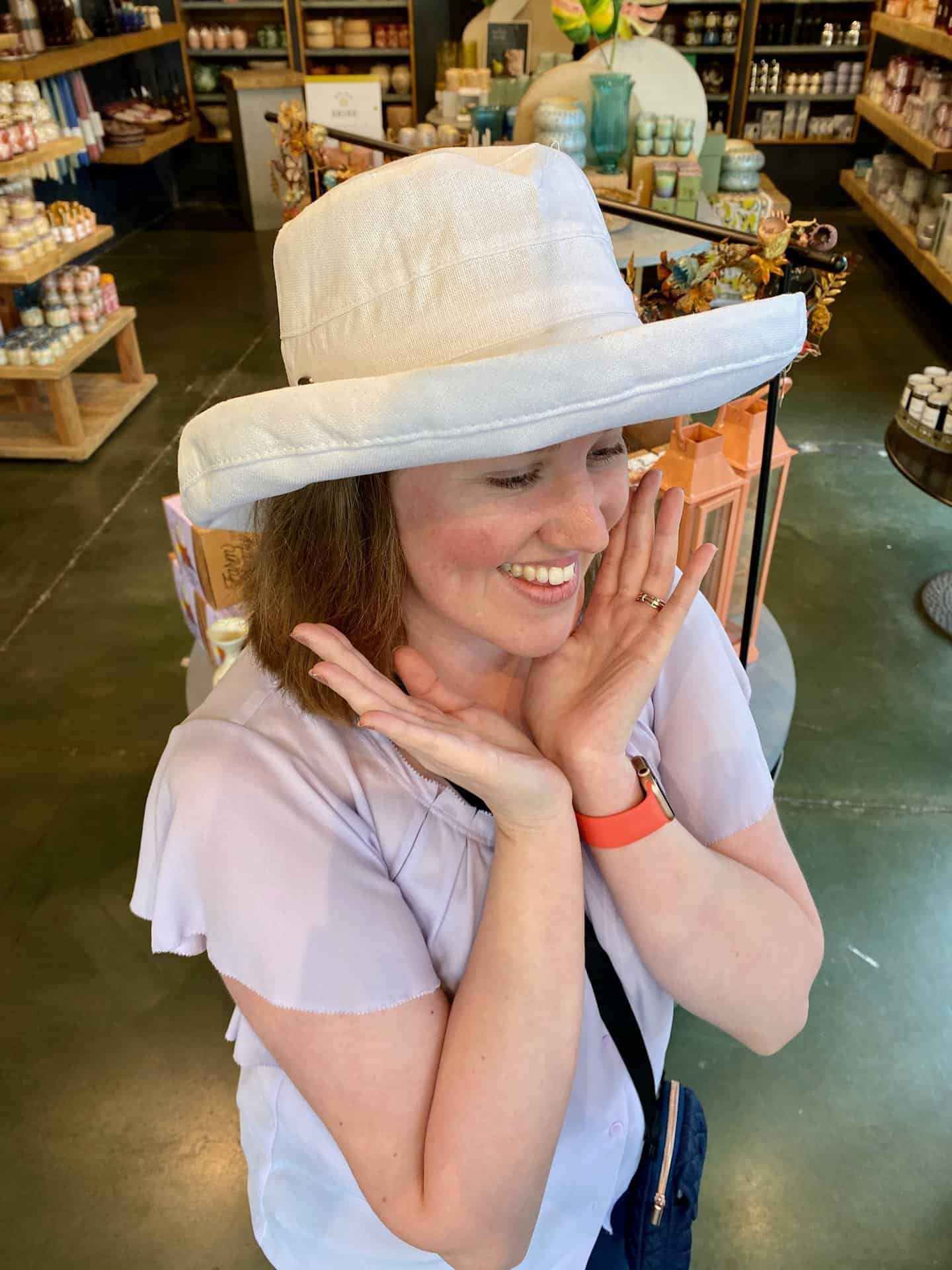 A person in a white sun hat and light purple blouse smiles and holds their hands to their face in a store with shelves of various products.