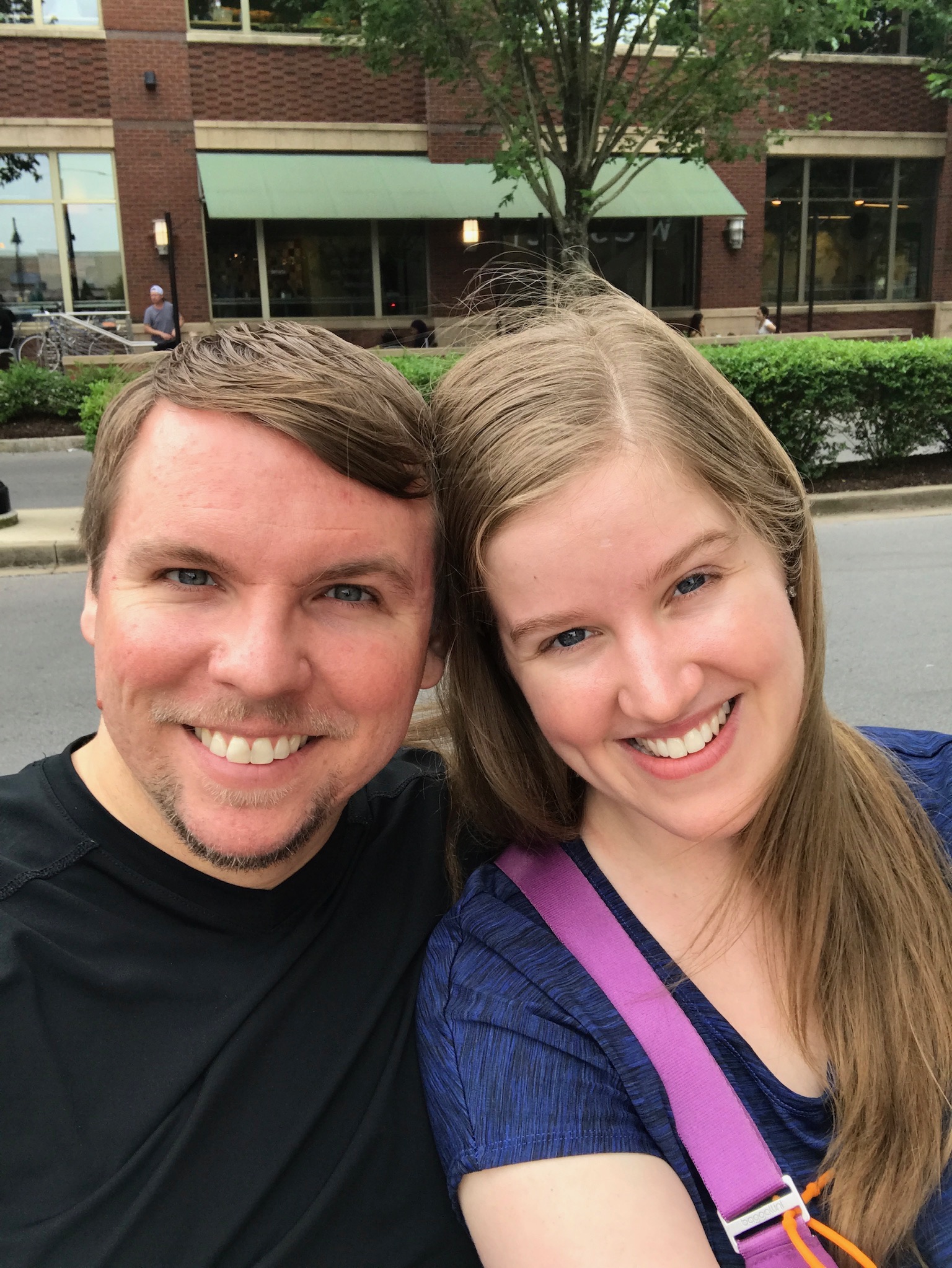A man and woman smiling together outdoors, sitting closely with a brick building, hedge, and tree in the background.
