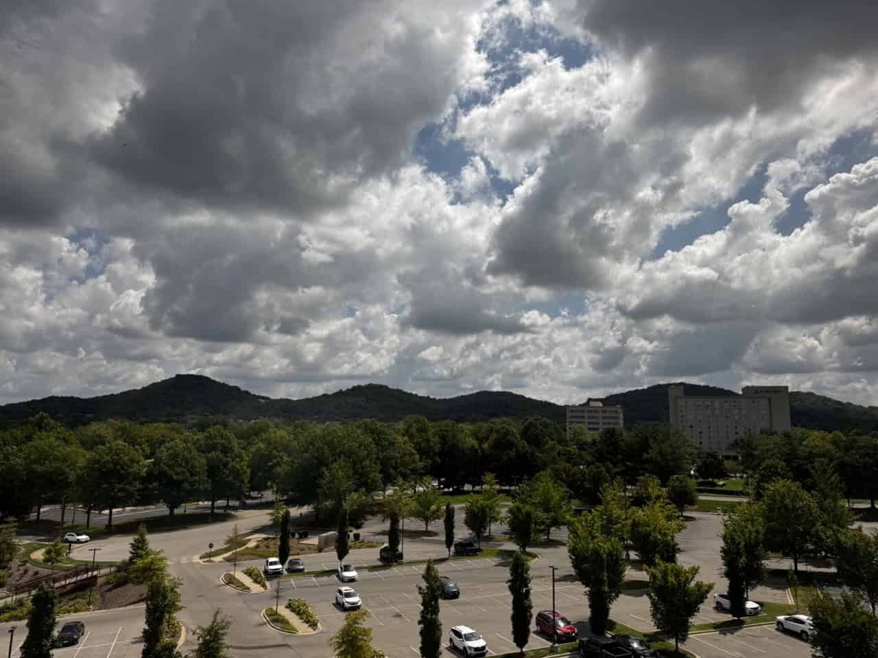 A parking lot filled with several cars stretches under a cloudy sky in Nashville. In the background, tree-covered hills and a few buildings complete the scene.