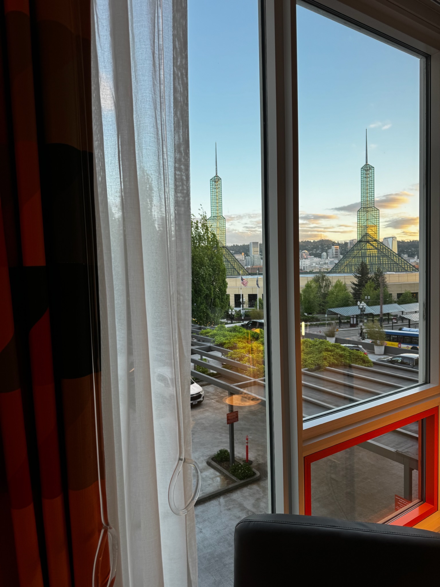 View through a window with white curtains showing a cityscape, including two tall glass spires, streets, greenery, and buildings under a blue sky with clouds.