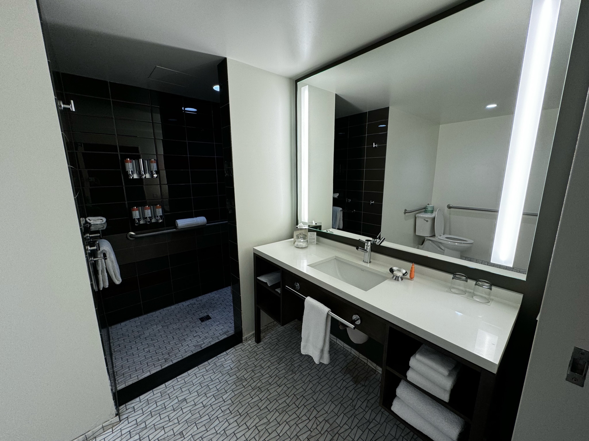 Modern hotel bathroom with large mirror, illuminated vanity, glass shower with black tiles, and towels neatly placed on open shelves under the sink. Toilet visible in background.