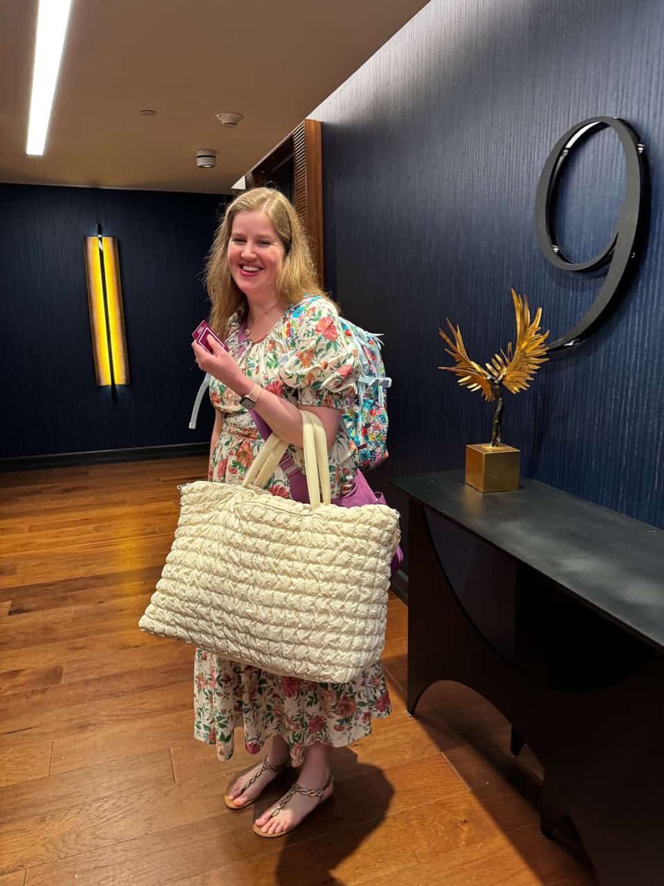 A woman in a floral dress stands in a Nashville hallway, holding a large woven bag and a drink cup, with the number 9 displayed on the wall behind her.
