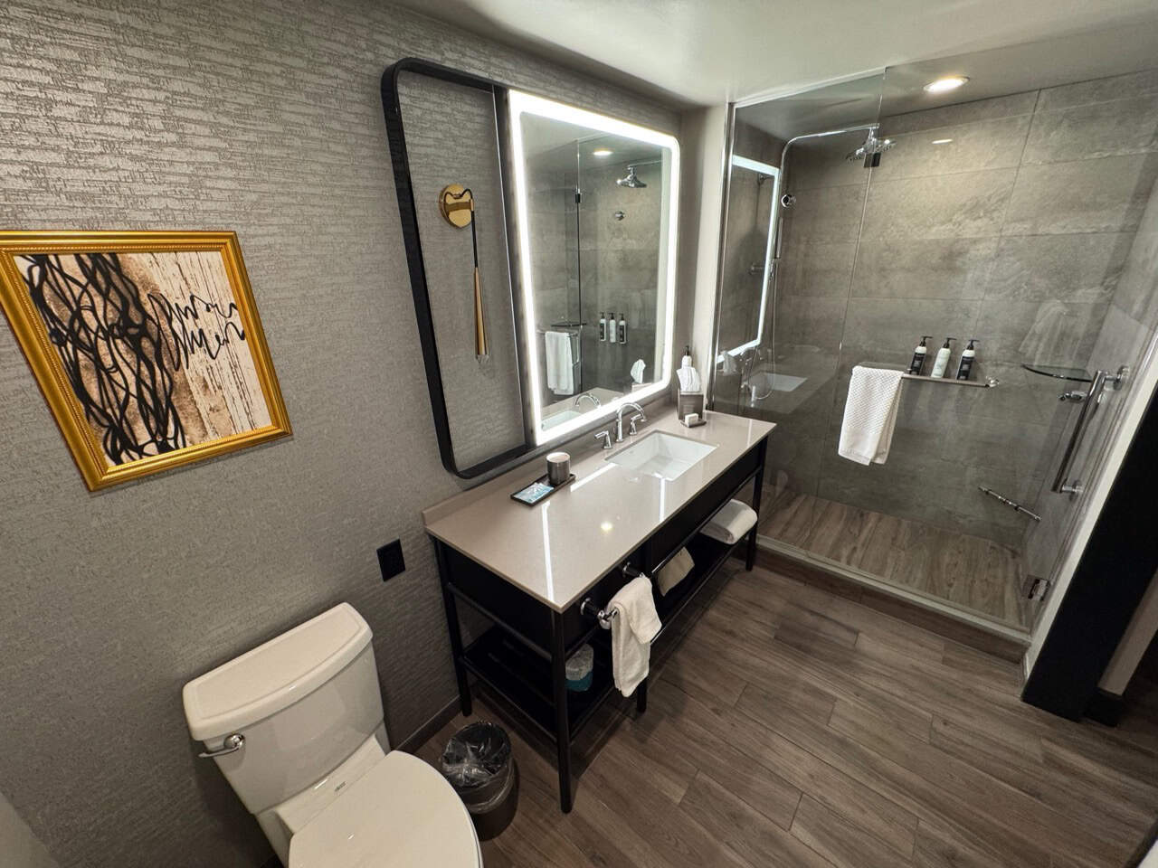 This modern Nashville bathroom features a large mirror and double sinks. A glass-enclosed shower is in view, while artwork adorns a textured wall above the toilet. The floor boasts wood-style tile, adding to the room's stylish appeal.