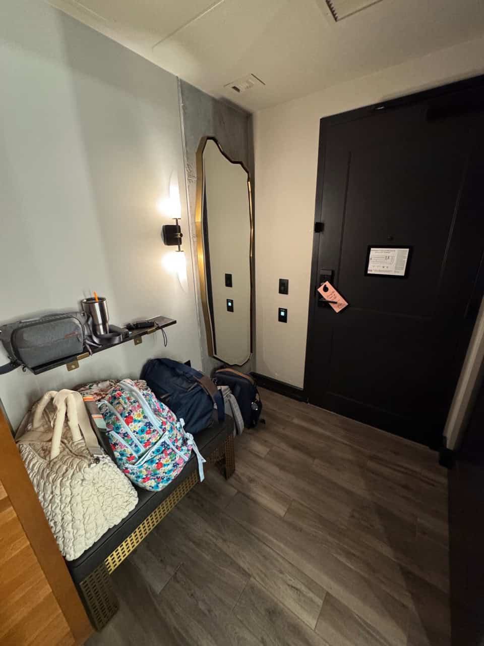 A Nashville-inspired hallway with a wooden floor showcases a bench adorned with a floral backpack, a beige throw, and a gray bag. A large mirror and a black door with a hanging sign complete this charming Southern scene.