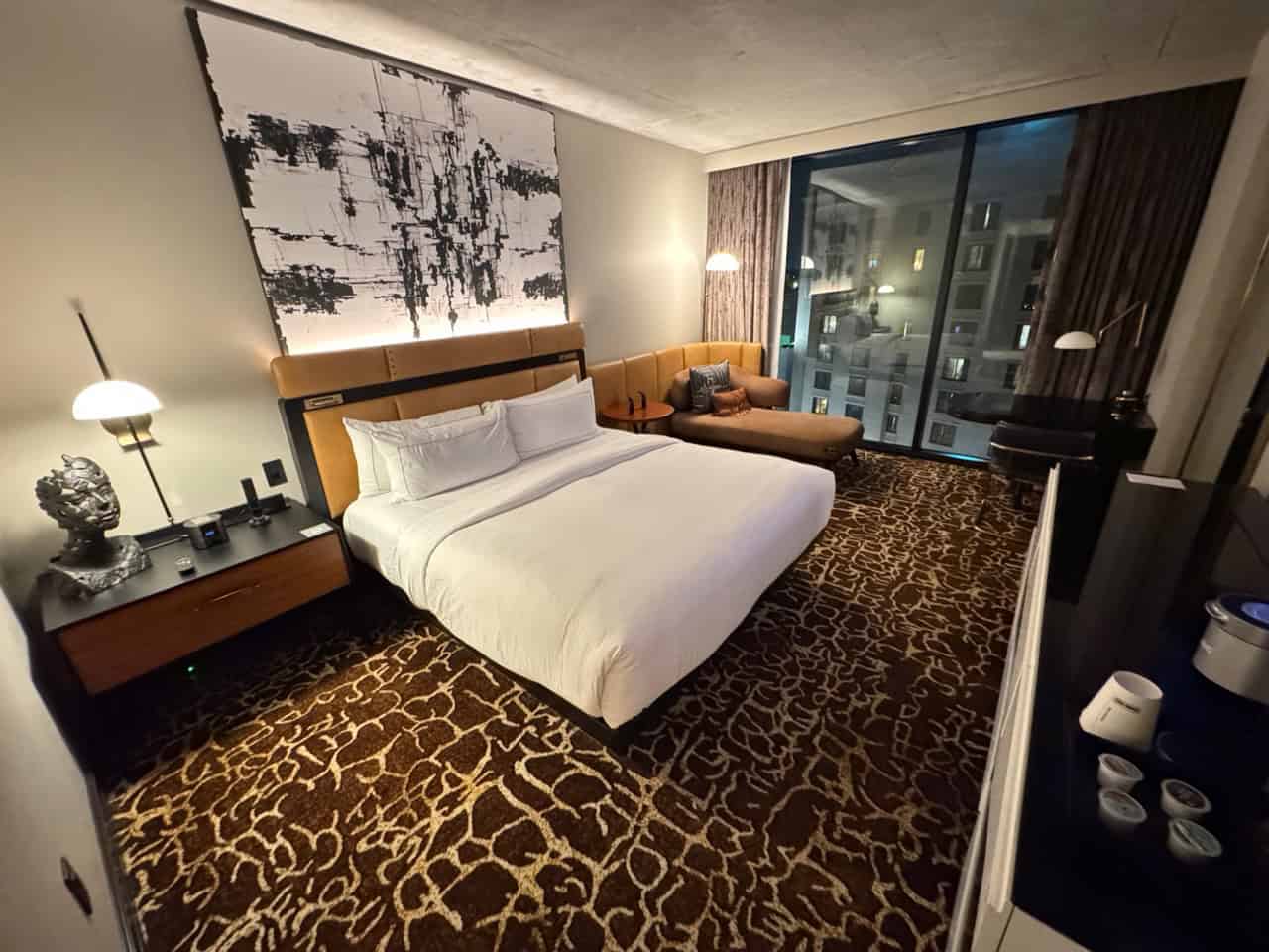 Hotel room with a bed, nightstand, abstract wall art, a cozy couch by the window, lamps, and a patterned carpet. Large window offers a stunning view of the Nashville cityscape.