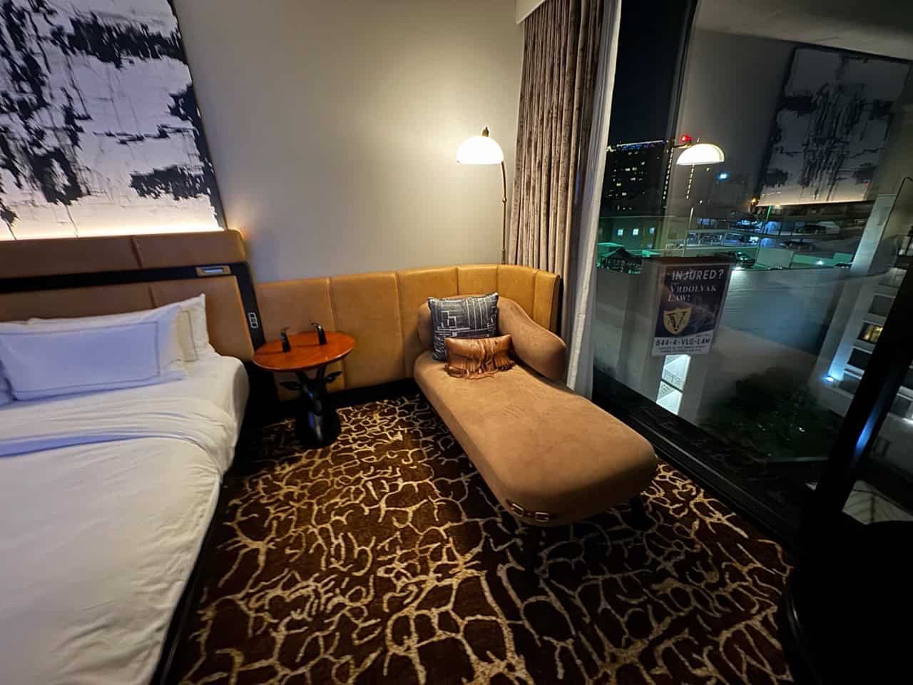 Hotel room with a king-size bed, a chaise lounge, and a floor lamp. Large window showcasing Nashville's night city view, abstract wall art, and patterned carpet.