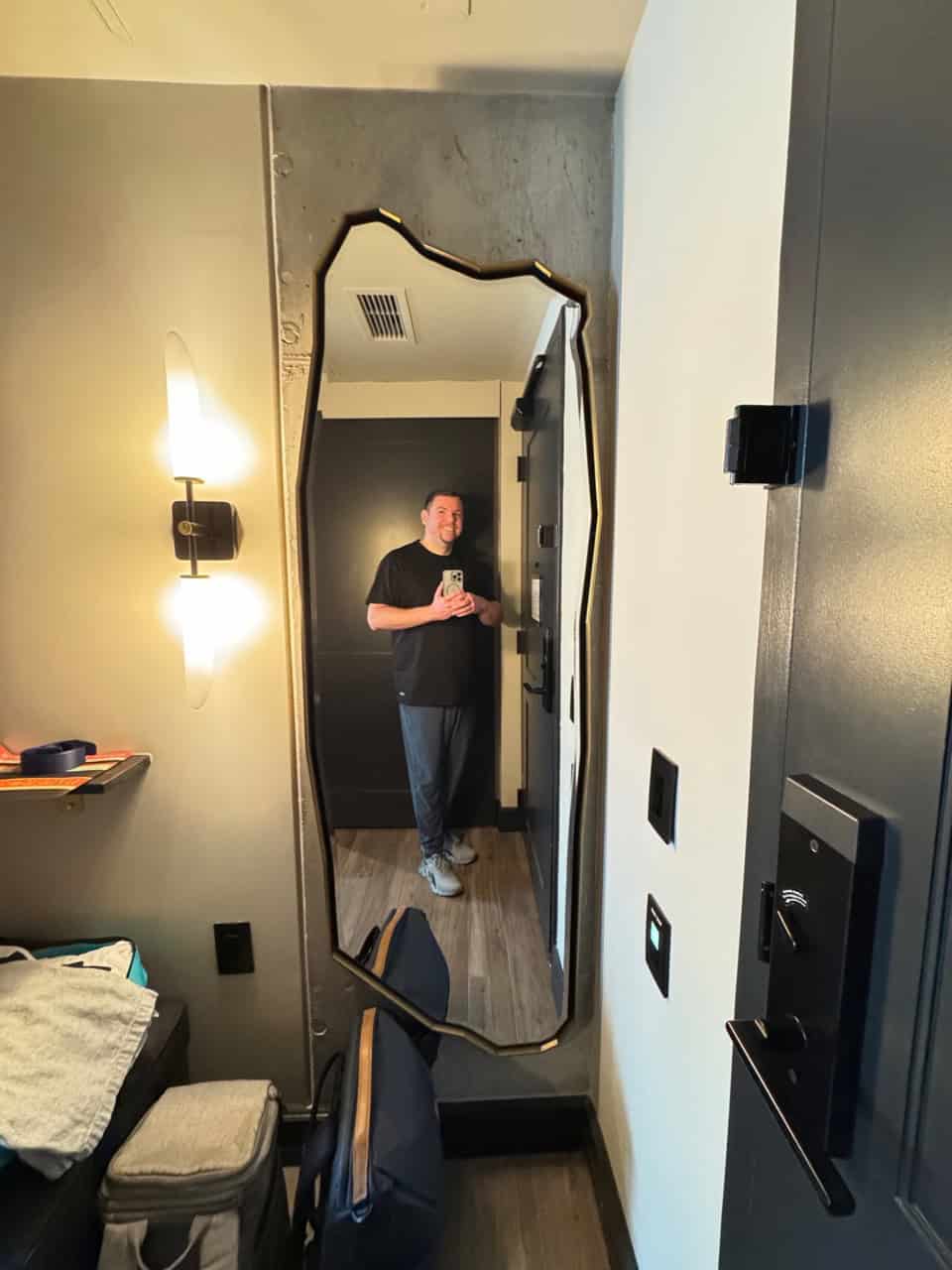 A person captures a selfie in a uniquely shaped mirror inside a modern Nashville room, featuring a wall light, textured walls, and a hint of their backpack.