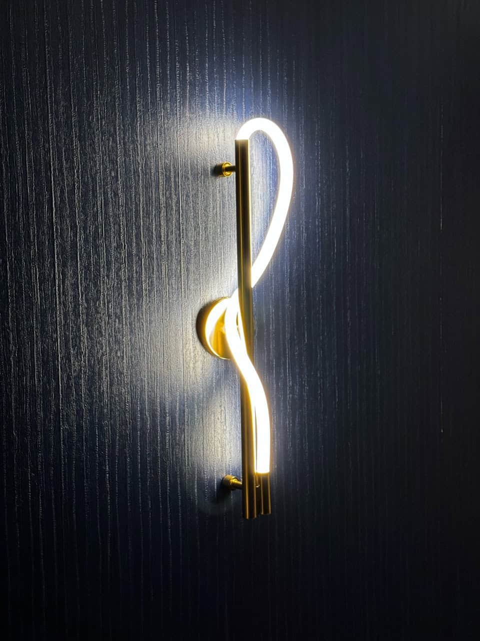 A modern wall-mounted LED light fixture, echoing Nashville's vibrant style, features a gold frame and looped design, illuminated brilliantly against a dark textured wall.