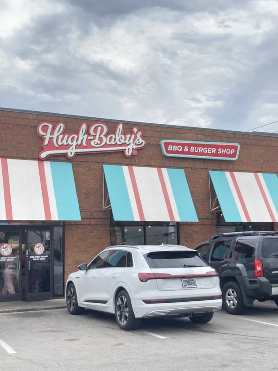 The exterior of Hugh Baby's, a Nashville brick BBQ and burger shop, boasts charming striped awnings. Under a cloudy sky, two cars are parked in front, ready for the culinary delights within.