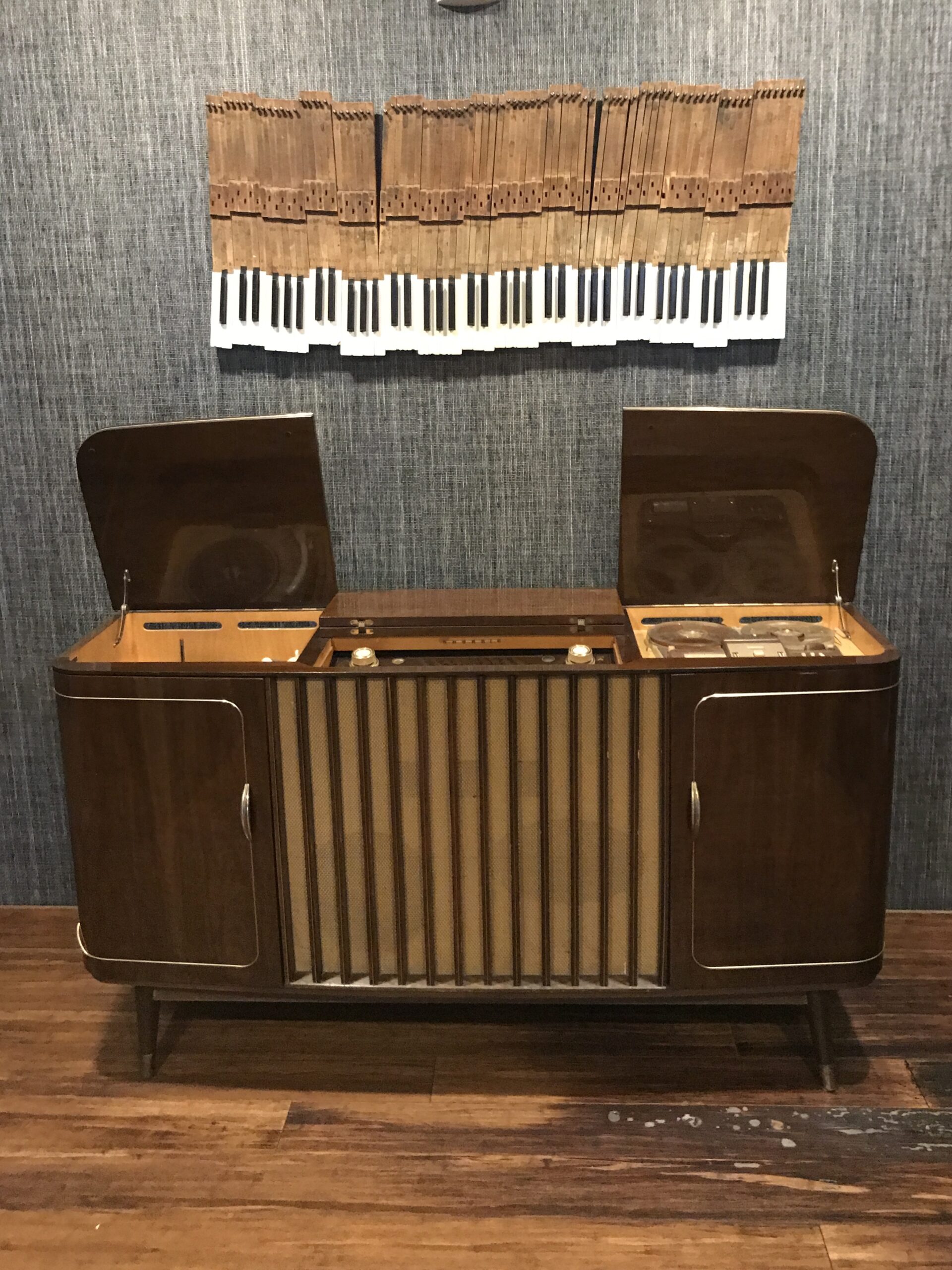 A vintage wooden stereo console with open lid compartments sits on a wood floor, beneath wall art depicting piano keys on a textured background.