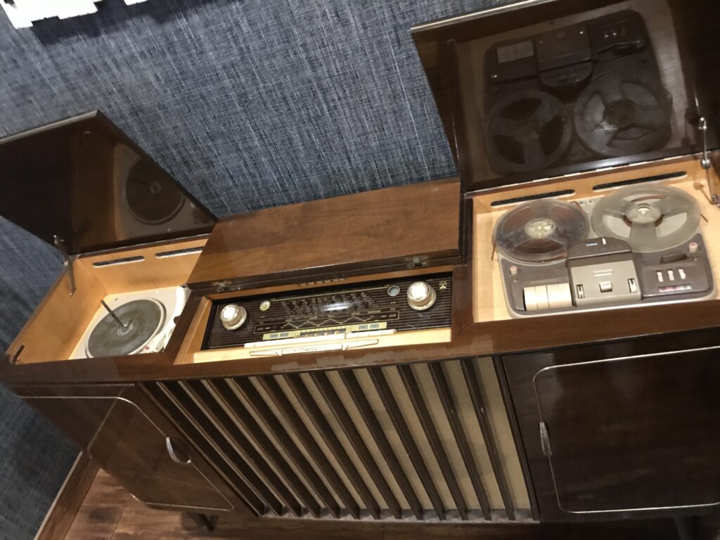 A vintage wooden audio console with a built-in record player, radio tuner, and reel-to-reel tape recorder, all with open lids.