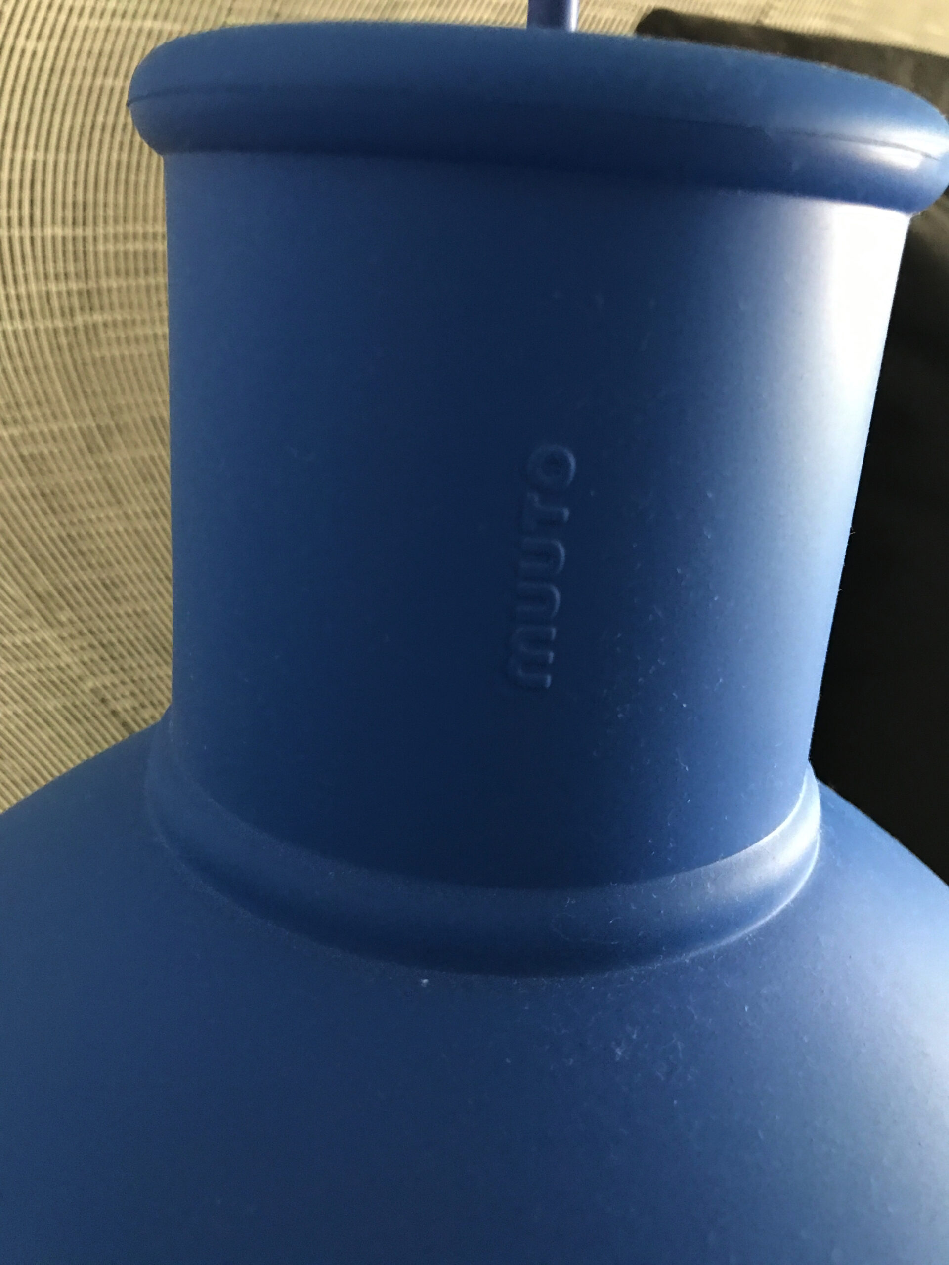Close-up of the neck of a matte blue Muuuto vase, with the brand name embossed vertically on the surface. A textured, light-colored background is partially visible.