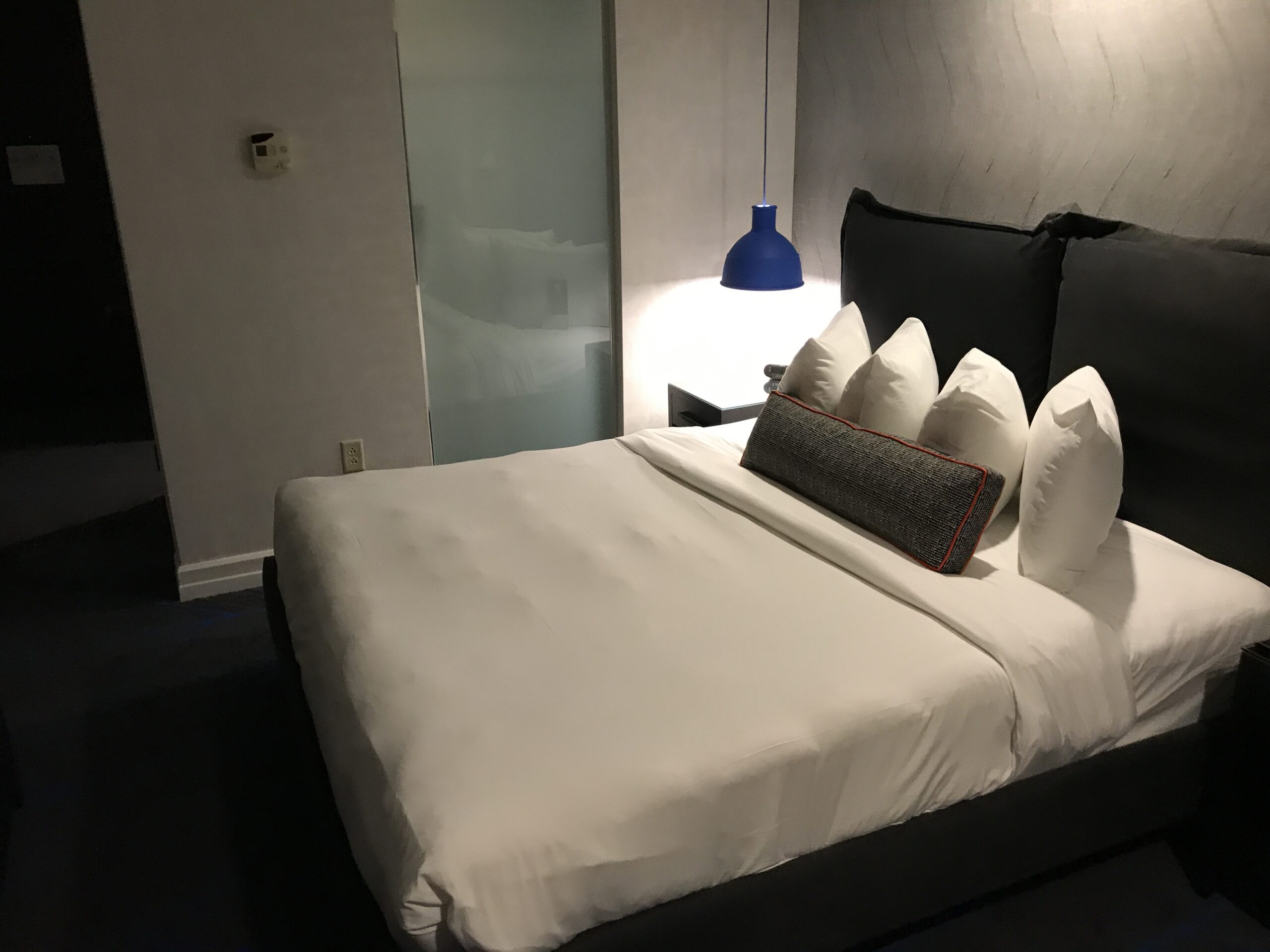 A neatly made bed with four white pillows and a bolster pillow in a modern hotel room, with a blue lamp on the nightstand and a frosted glass wall in the background.
