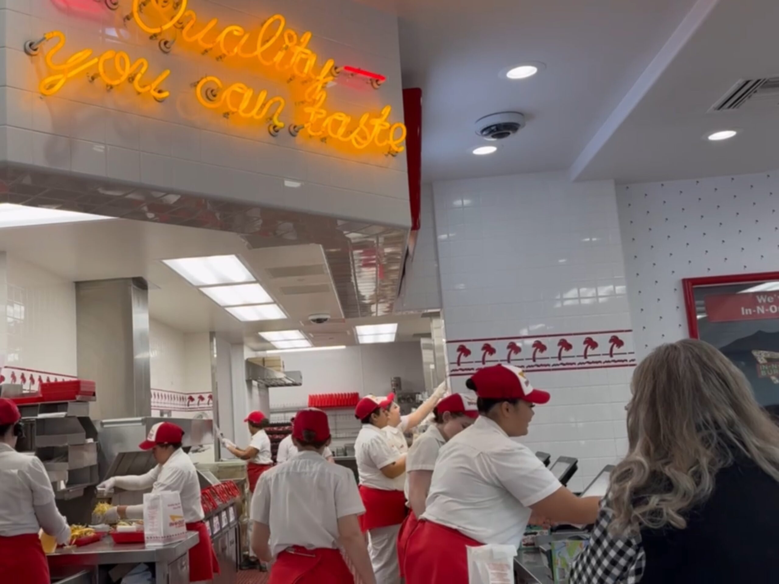 Several employees in red hats and white uniforms work behind the counter at a fast food restaurant as a customer waits. A neon sign reads "Quality you can taste.