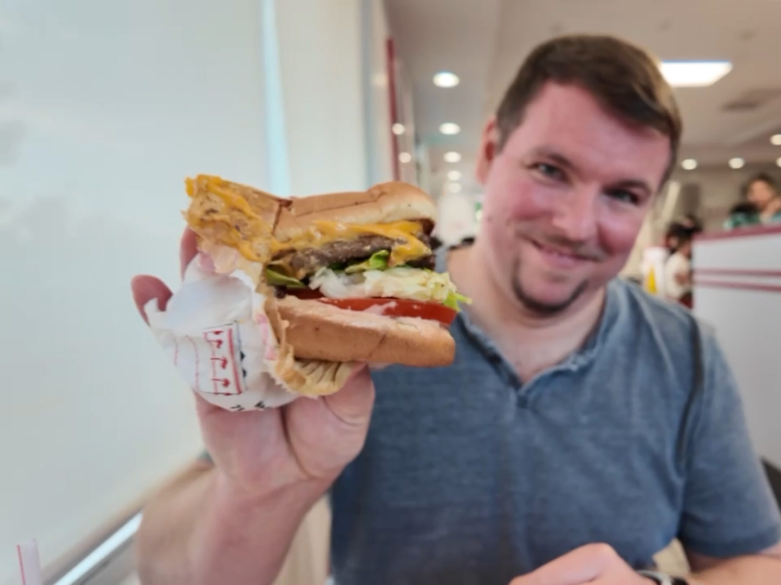 A man in a blue shirt holds up a cheeseburger with lettuce, tomato, and cheese inside a fast food restaurant.