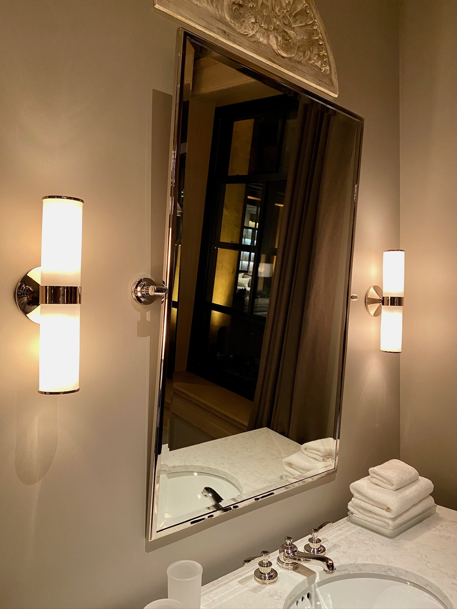 A bathroom mirror above a marble sink, flanked by two wall-mounted lights, with neatly stacked white towels and cups on the countertop, brings a hint of Nashville charm to this stylish space.