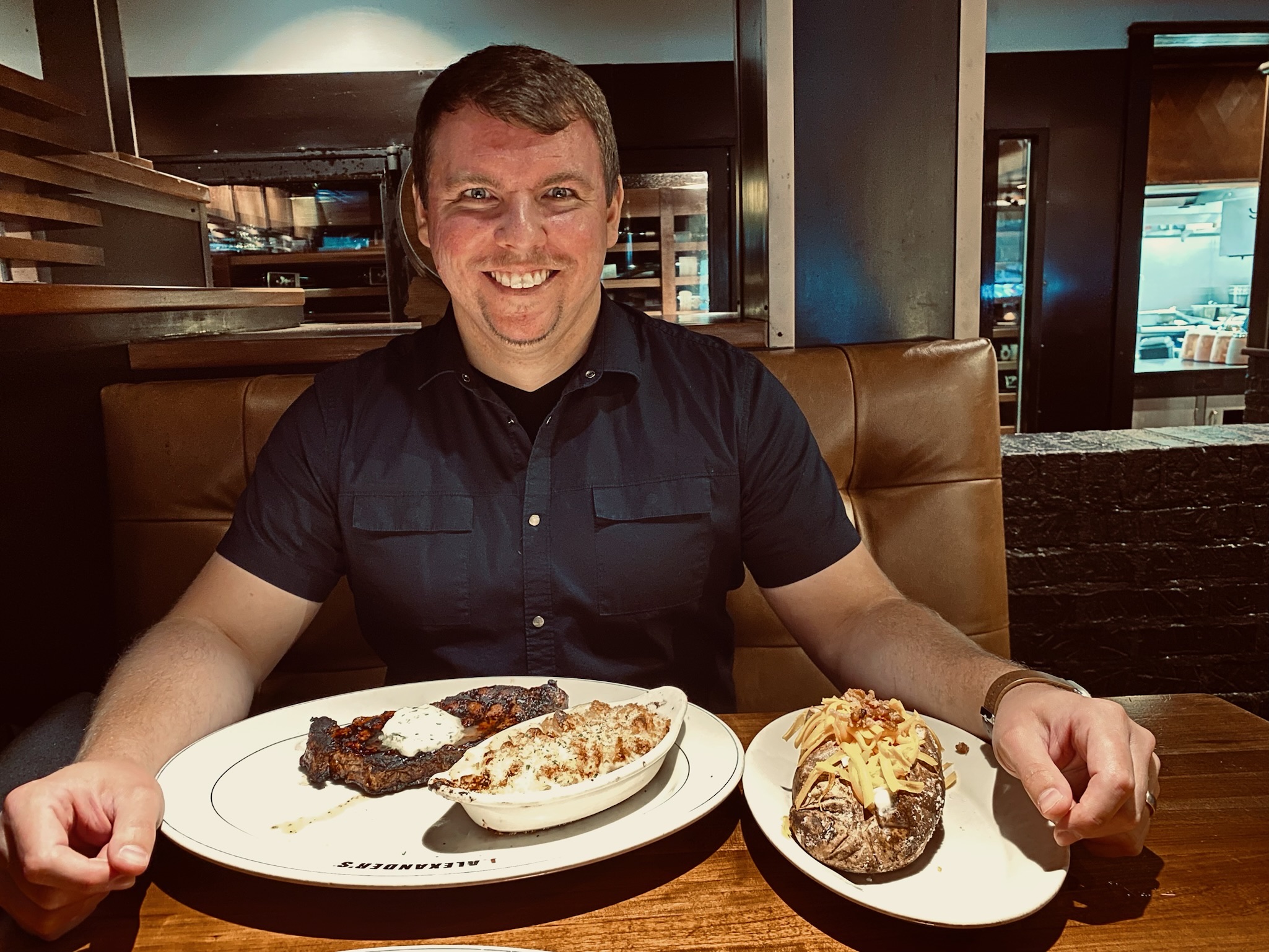 A man sitting at a restaurant table smiles at the camera with three dishes in front of him: steak, a baked potato with toppings, and a casserole.