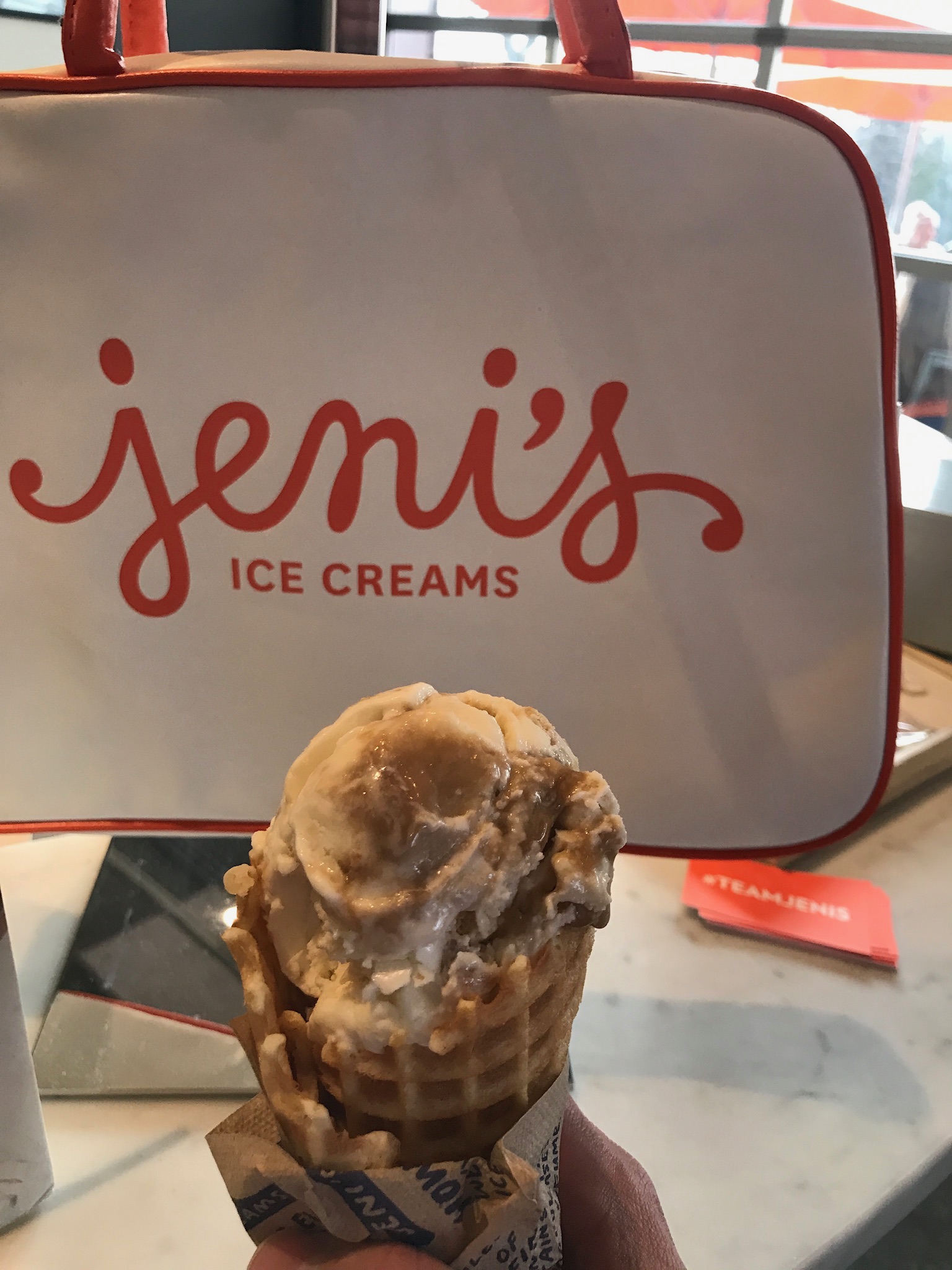 A hand holds an ice cream cone in front of a Jeni's Ice Creams branded cooler bag on a marble countertop.