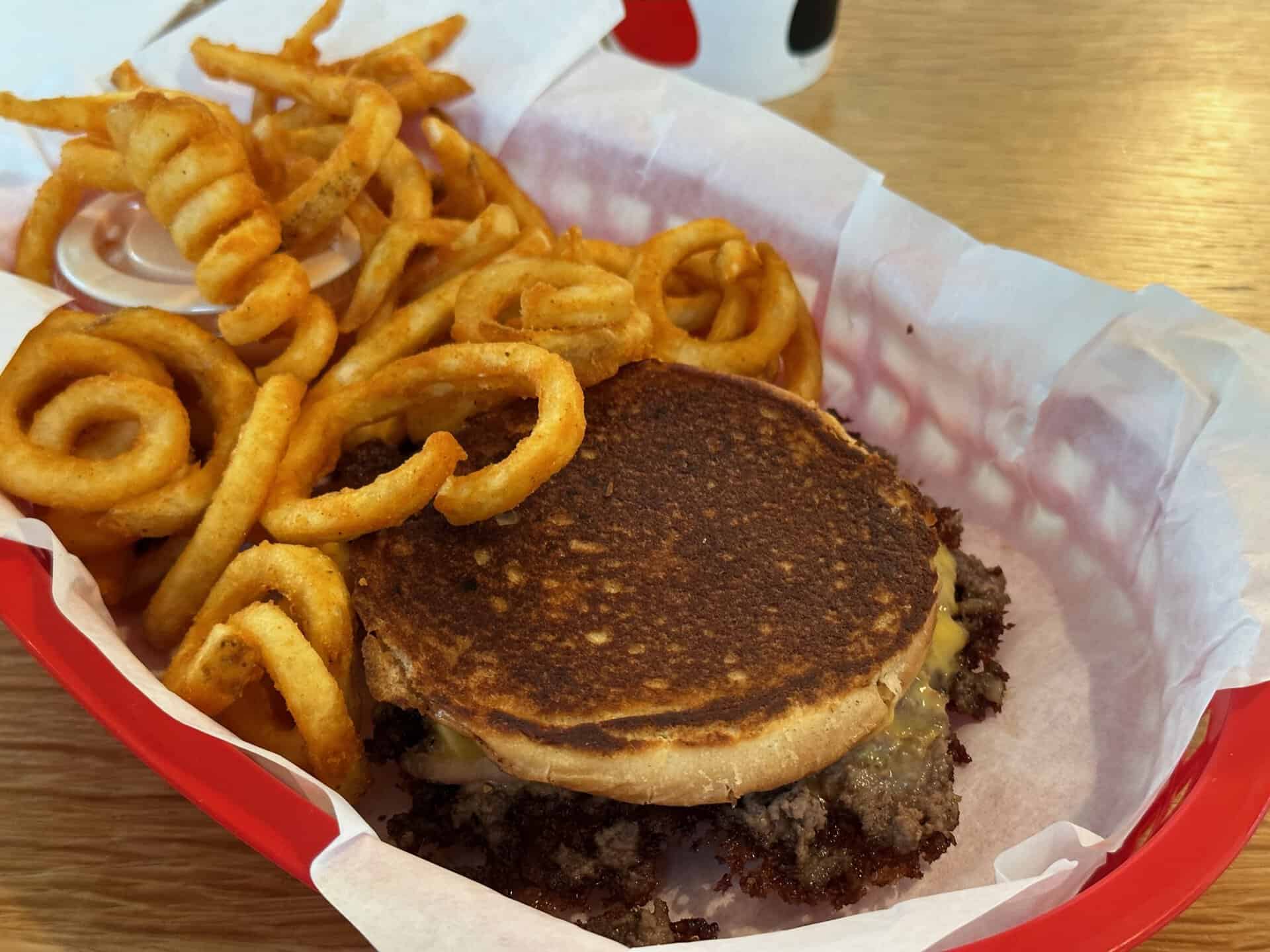 A Nashville twist on classic comfort: Enjoy a grilled cheese sandwich stacked with a juicy beef patty, served up with crispy curly fries in a red basket lined with white paper.