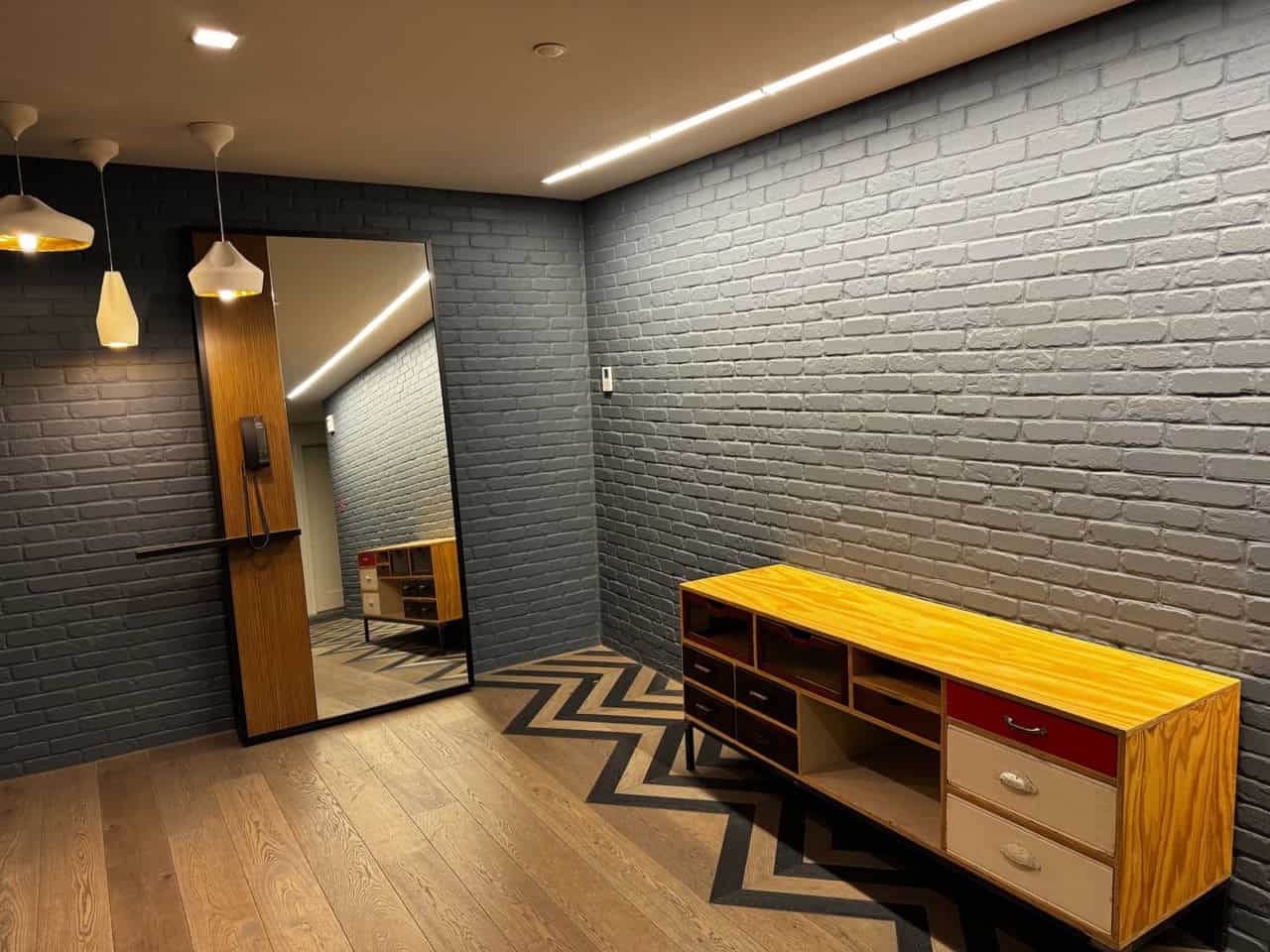 Room with gray brick walls, wooden floor, and a zigzag pattern rug captures the Nashville charm. A large mirror reflects the warmth of a wooden cabinet with drawers, while three white pendant lights complete the cozy ensemble.