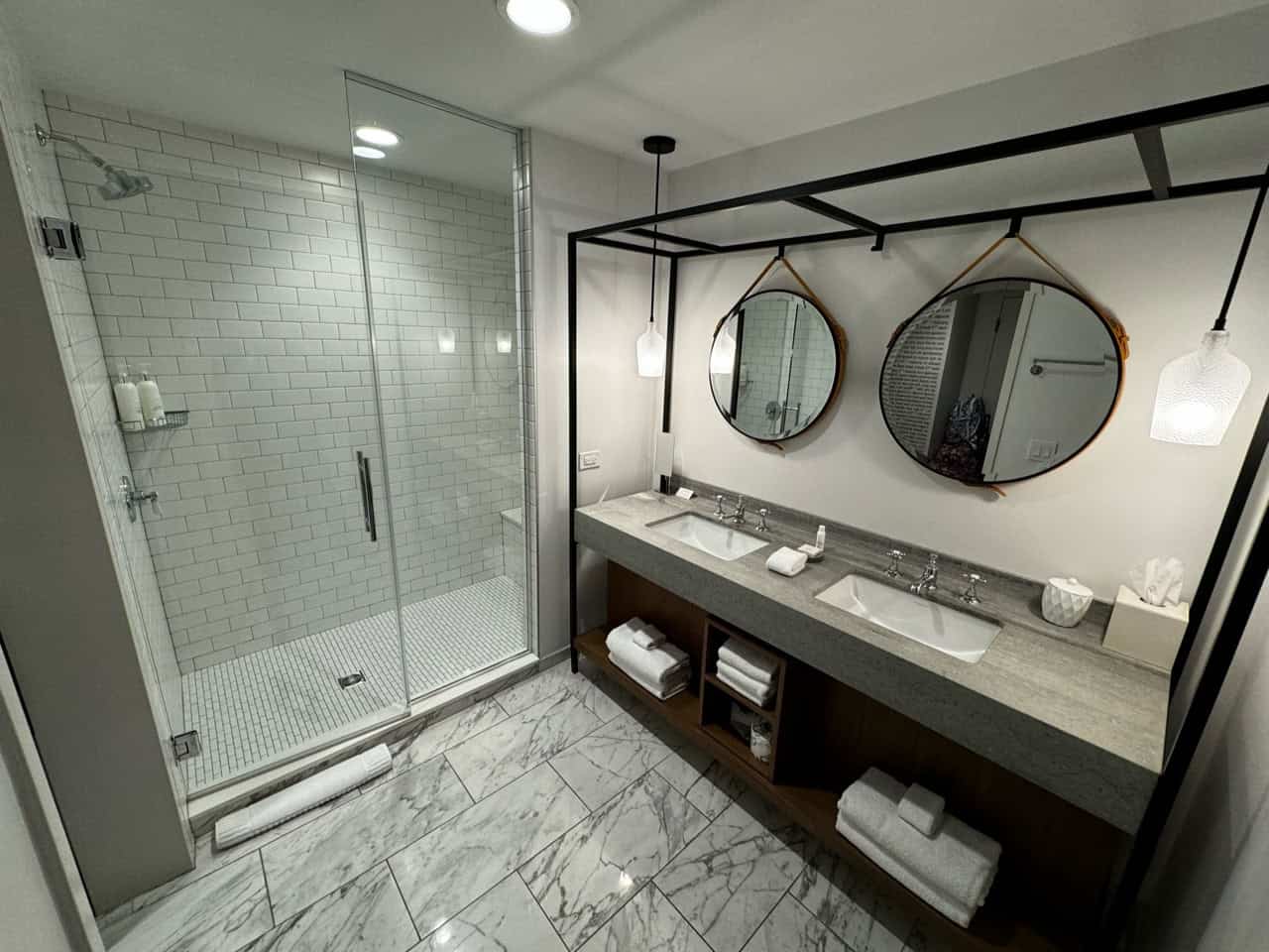 A Nashville-inspired modern bathroom features a glass shower, two round mirrors, and dual sinks on a concrete countertop. Marble floor tiles add elegance, while towels and toiletries are neatly organized below the counter.