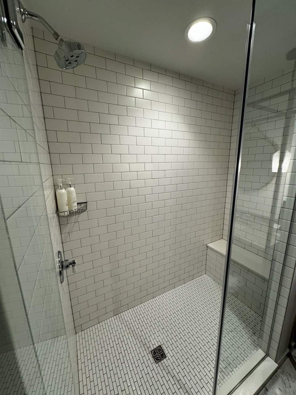 A modern Nashville shower with white subway tile walls and a glass door, featuring two shampoo bottles on a corner shelf and a small built-in bench.