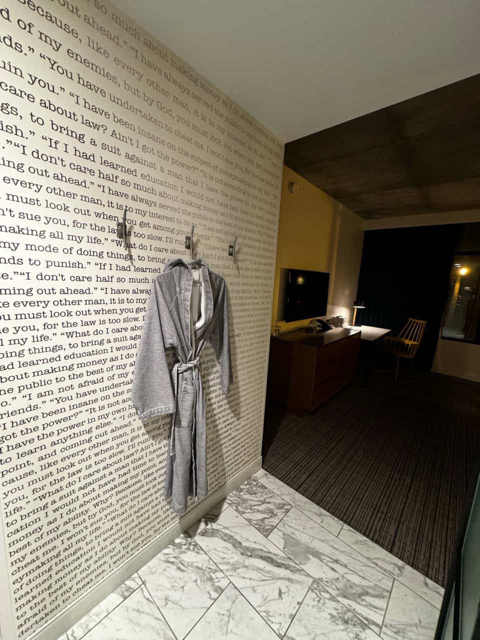 A hallway with a robe hanging on a wall covered in text evokes a cozy southern charm. In the dimly lit room beyond, a Nashville-inspired workspace features a desk with a chair, monitor, and inviting lamp.