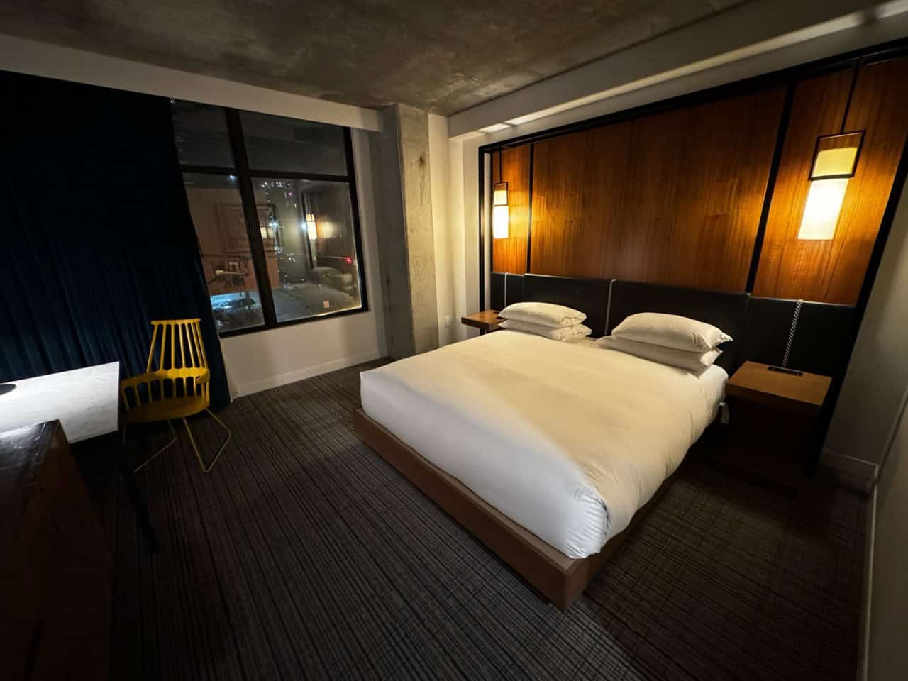 A modern Nashville hotel room featuring a large bed, two bedside tables with lamps, a sleek desk with a chair, and a window adorned with dark curtains.