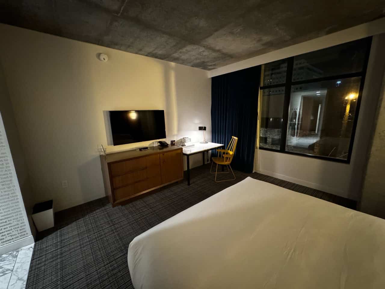 A modern Nashville hotel room features a bed, wall-mounted TV, wooden desk, chair, and large window. The decor is minimalistic with a neutral color palette, blending the vibrant spirit of the city with serene comfort.
